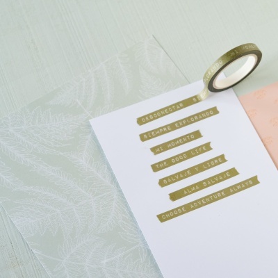 Fita adesiva dourada com frases sobre papel branco e papéis translúcidos decorativos ao lado.