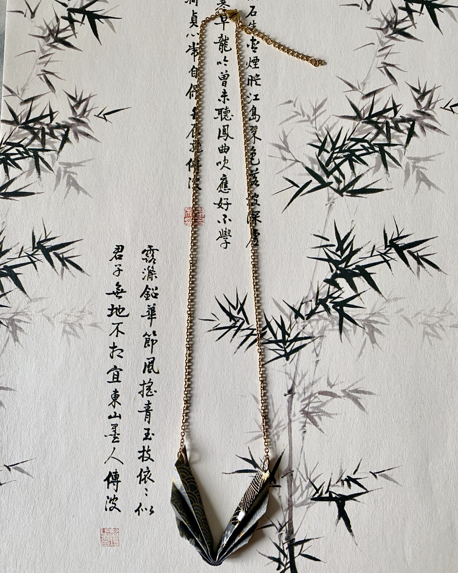 Colar Niki Colar dourado com pingente origami em papel com padrão preto e dourado sobre papel com caligrafia e bambu