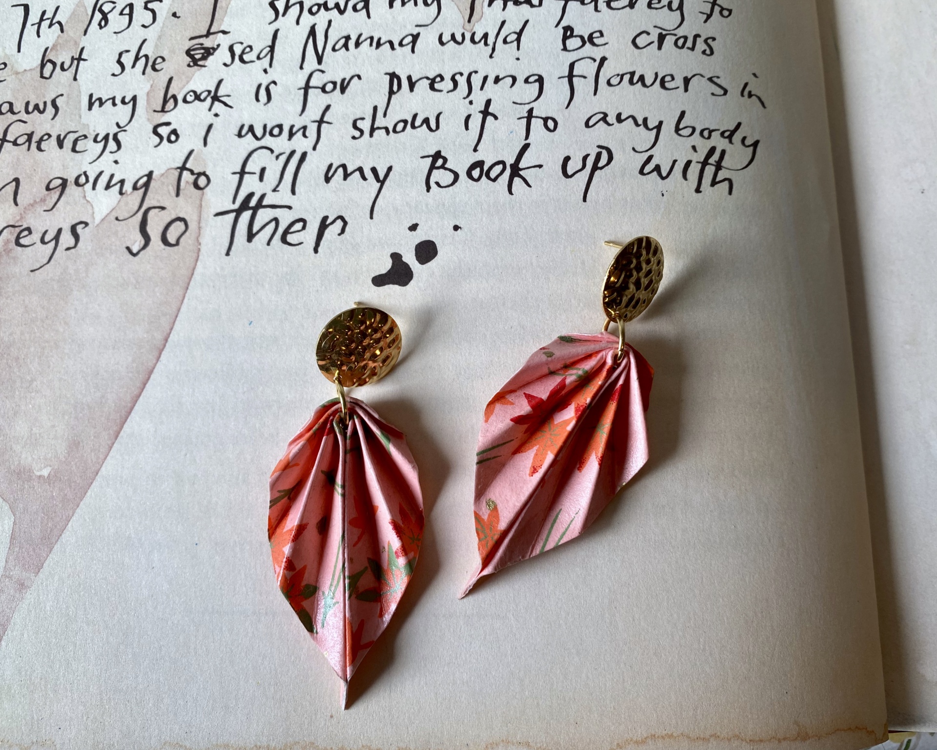 Brincos Folha Brincos com base dourada e pendente de papel rosa com padrão floral, sobre página de livro manuscrito.