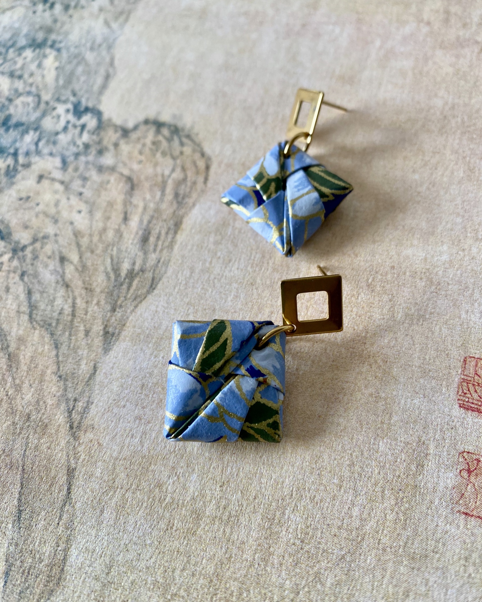 Brincos Sonobe Brincos dourados com elemento azul e verde dobrado, sobre superfície creme com desenho floral