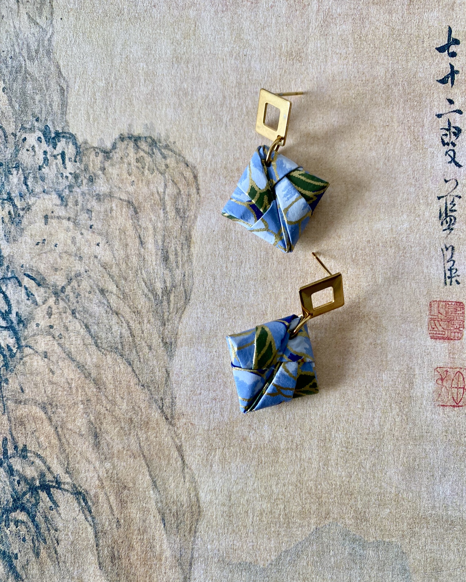 Brincos Sonobe Brincos quadrados em tecido azul com padrão floral verde e encaixes dourados sobre papel com texto oriental.