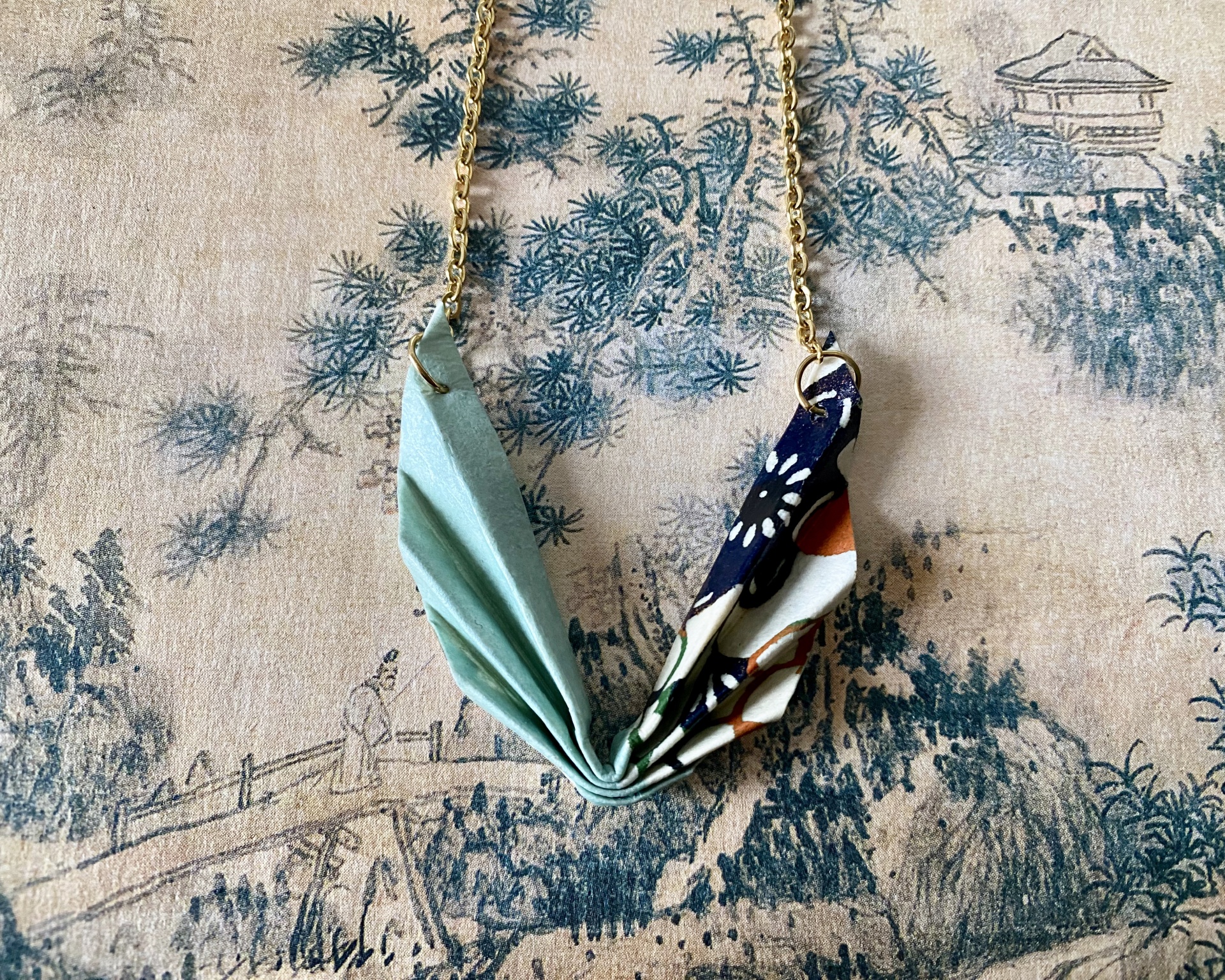 Colar Niki Colar com pendentes de tecido azul claro e floral em fundo estampado de paisagem.