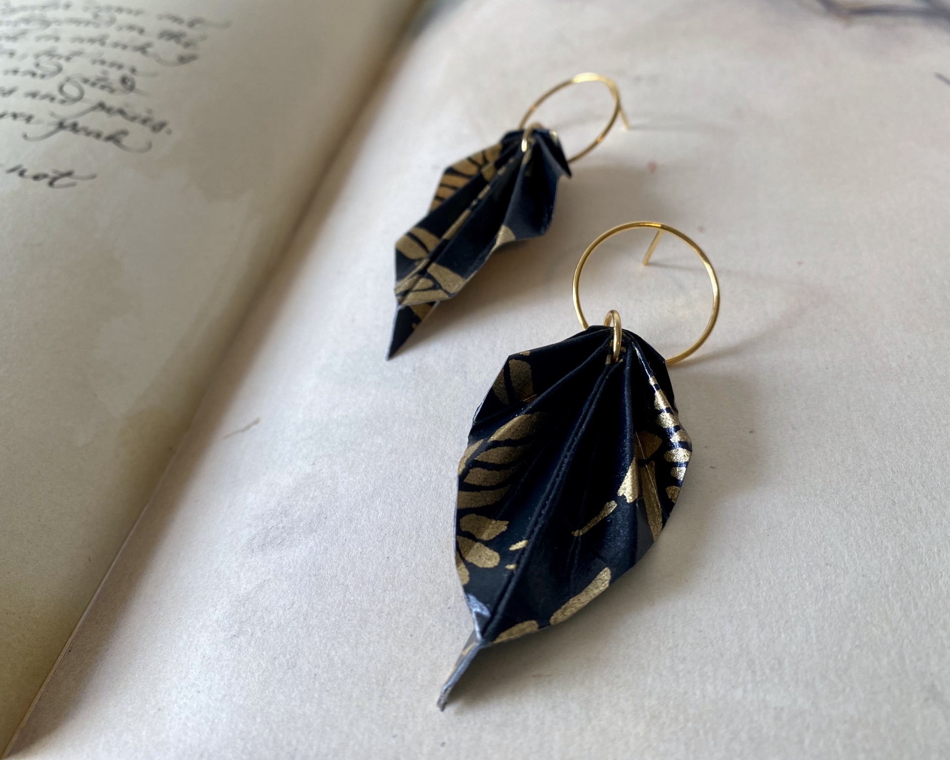 Brincos Folha Brincos com pendentes de tecido preto e dourado em formato de folha sobre superfície branca junto a livro aberto
