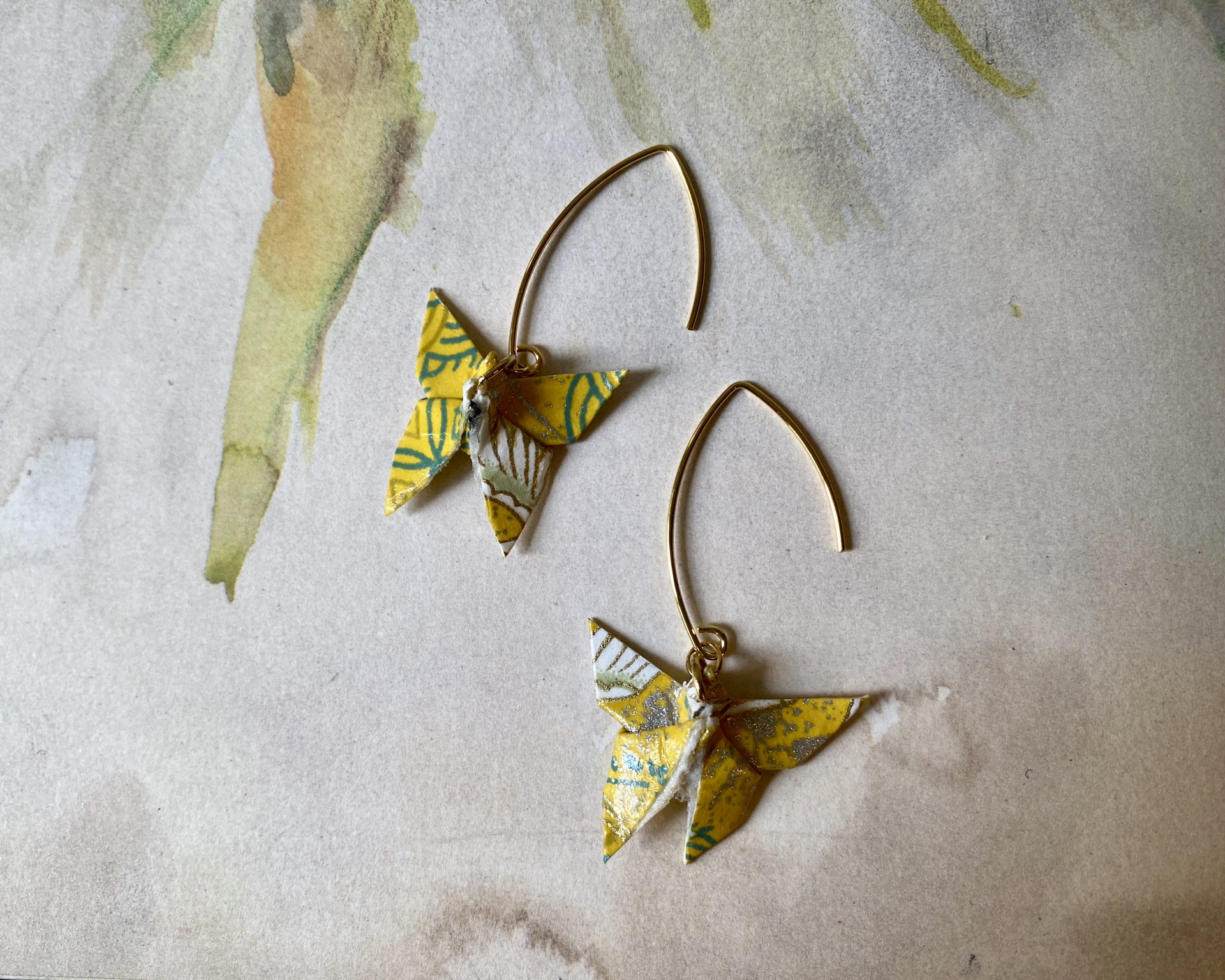 Brincos Borboleta Brincos com pendente origami amarelo e argola dourada sobre fundo de papel aquarelado