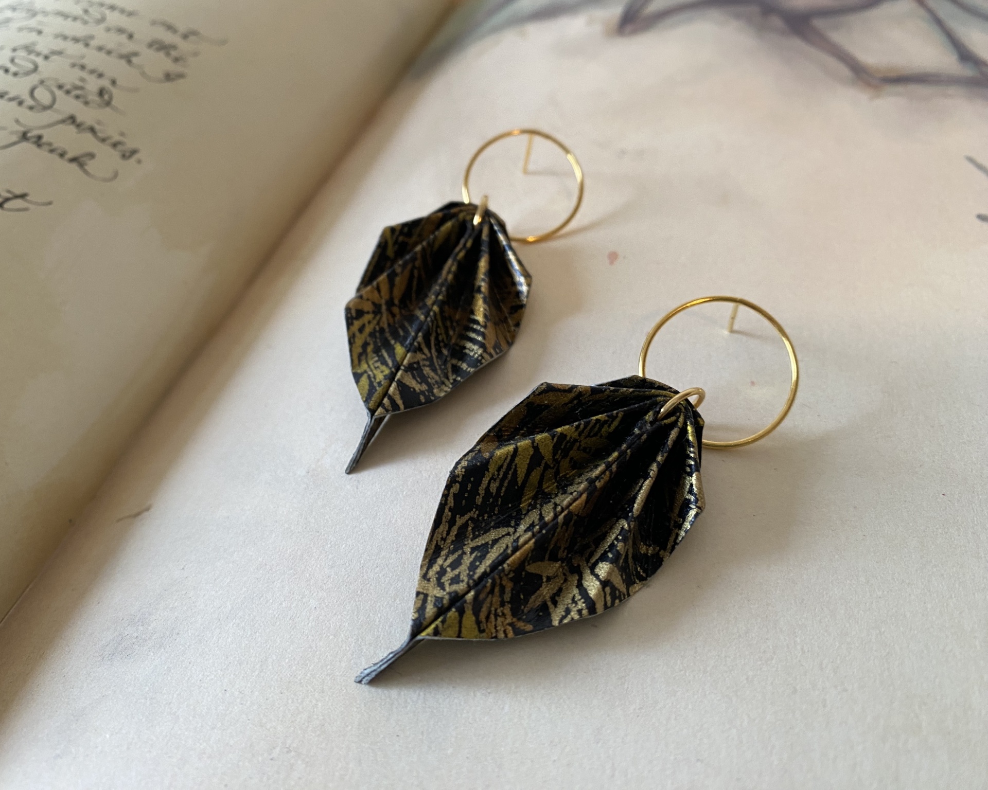 Brincos Folha Brincos com argolas douradas e pendentes em forma de folhas preto e dourado sobre página de livro aberto