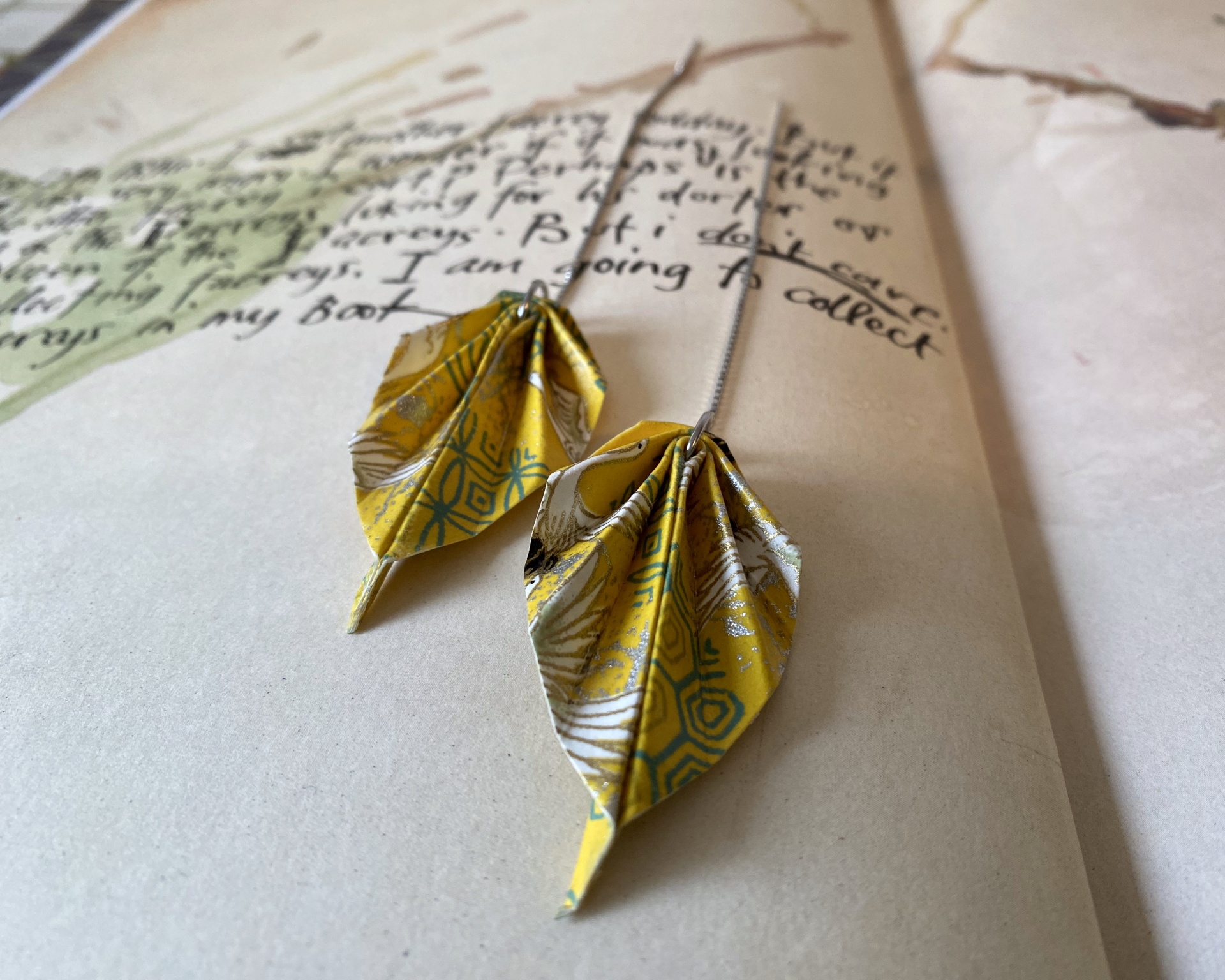 Brincos Folha Brincos pendentes de origami amarelos com padrões, sobre papel com texto manuscrito.