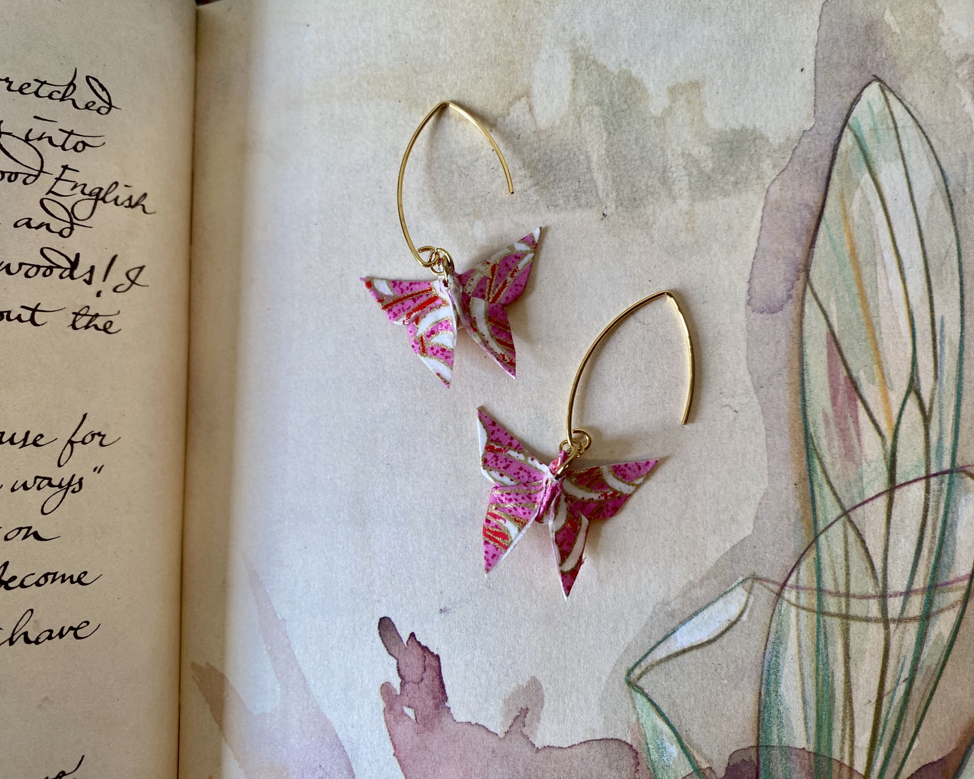 Brincos Borboleta Brincos de borboleta rosa e branco com arame dourado sobre livro aberto.