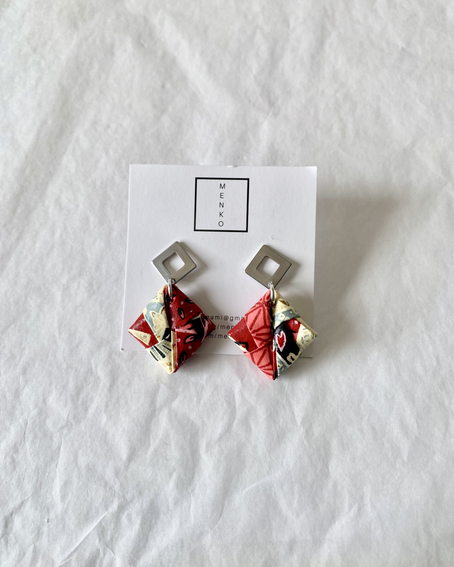 Brincos Sonobe Brincos geométricos em metal prateado e tecido colorido em cartão branco com a marca MENKO