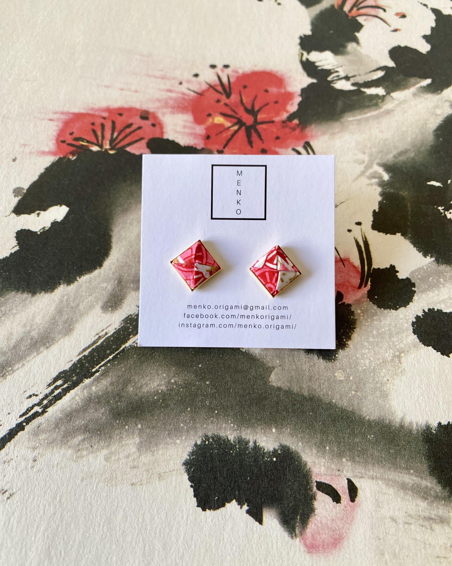 Brincos Menko Clássico Brincos de origami vermelhos e brancos montados em cartão branco com logo MENKO, sobre pintura floral