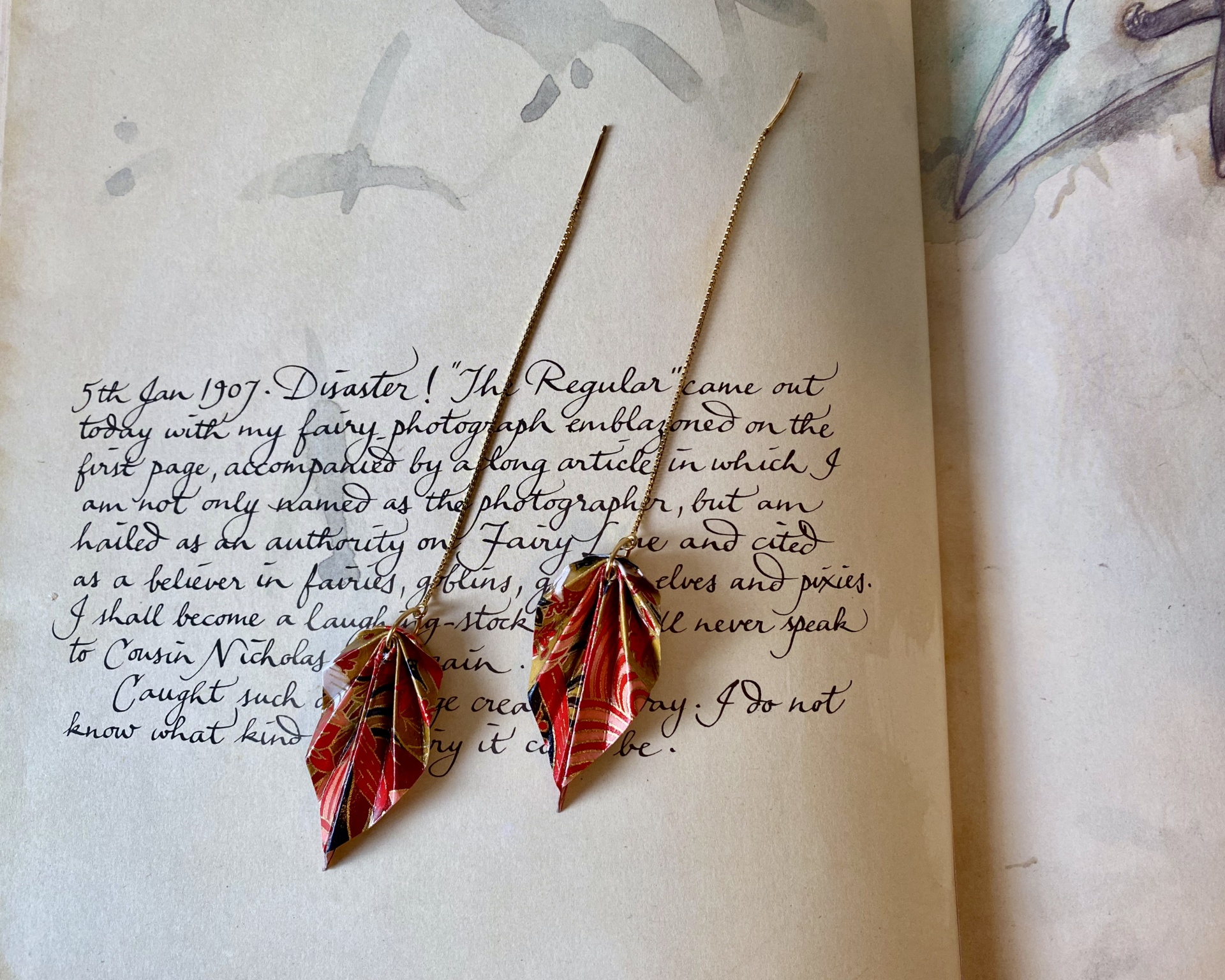 Brincos Folha Brincos dourados com folhas vermelhas sobre página manuscrita