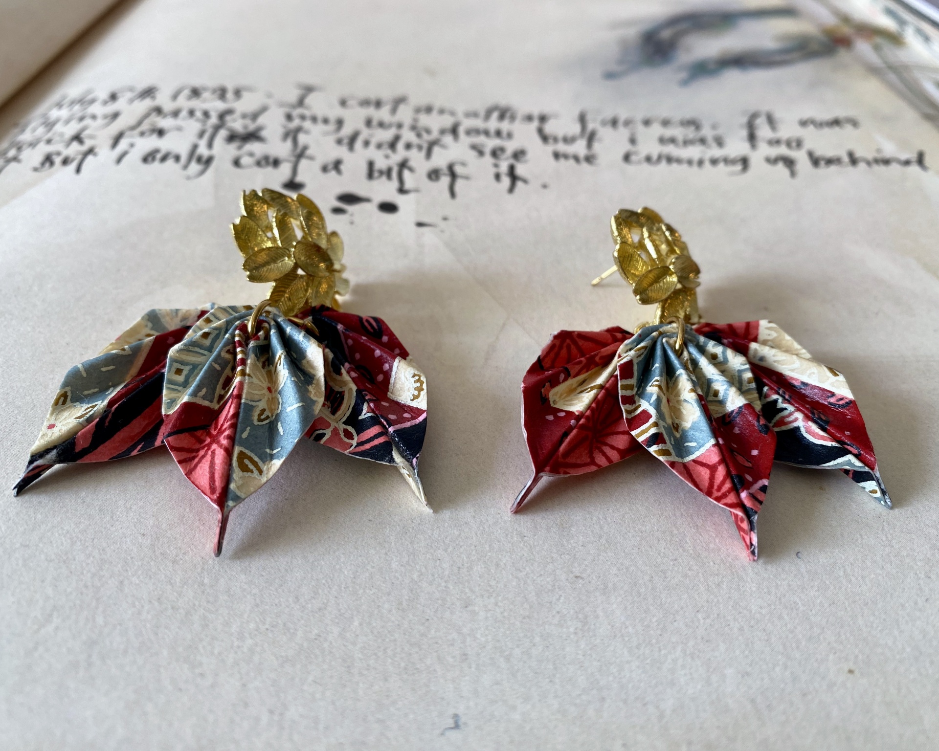 Brincos Folha Brincos dourados com pendentes de tecido estampado colorido sobre papel manuscrito.