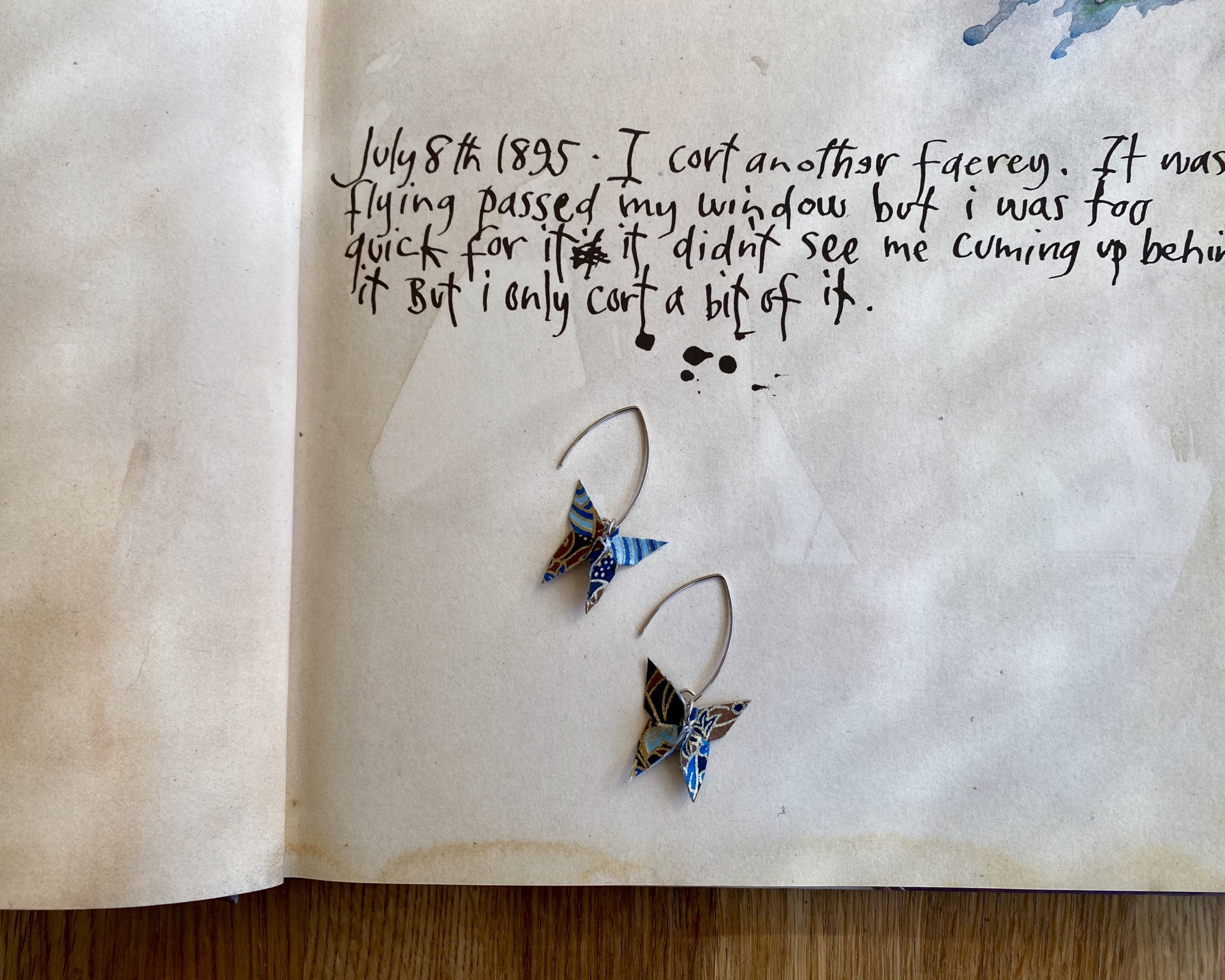 Brincos Borboleta Página manuscrita com dois brincos pendurados em forma de asas de borboleta azuis e castanhas.