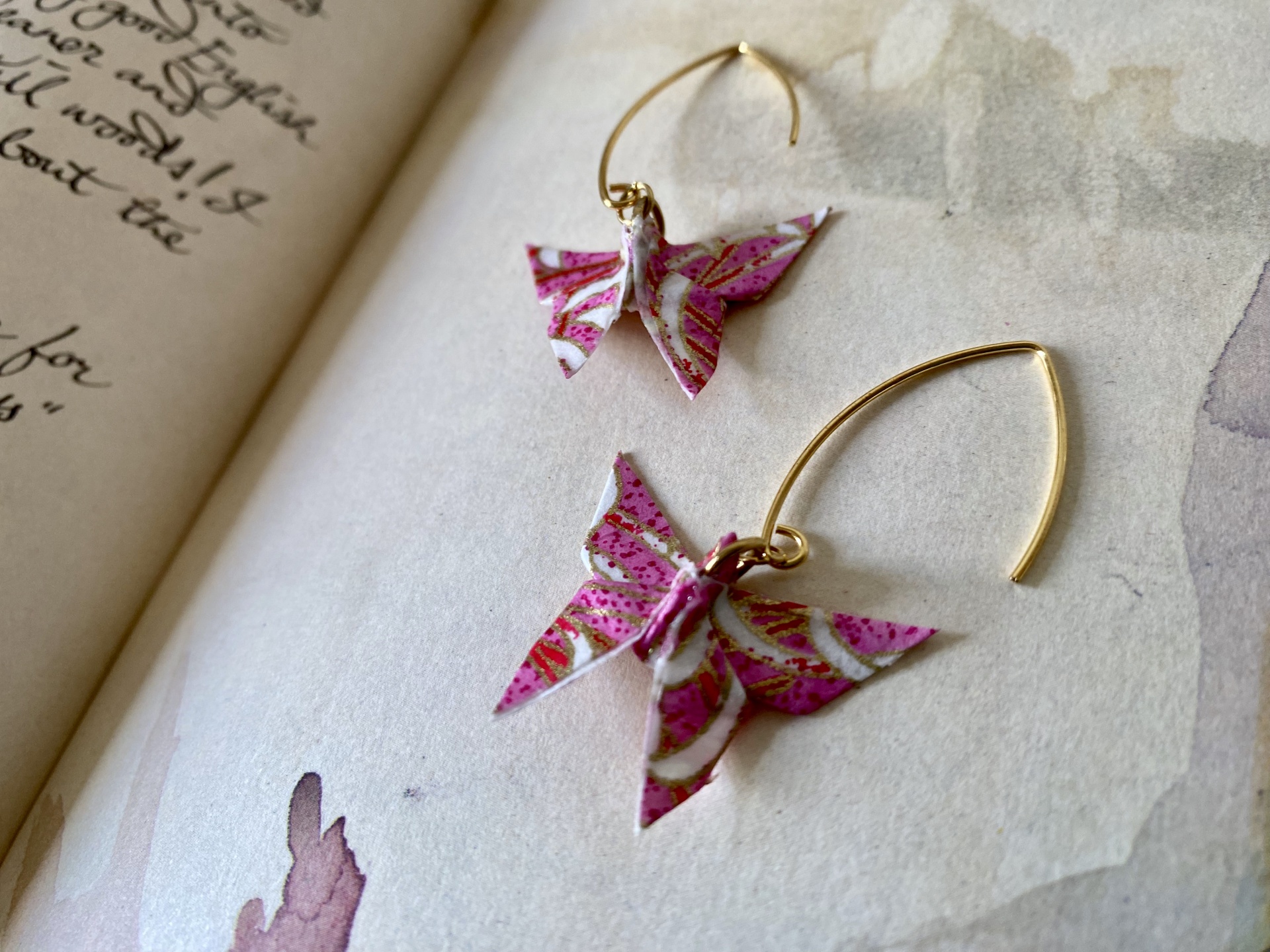 Brincos Borboleta Brincos dourados com borboletas cor-de-rosa com detalhes brancos e dourados sobre papel com texto manuscrito