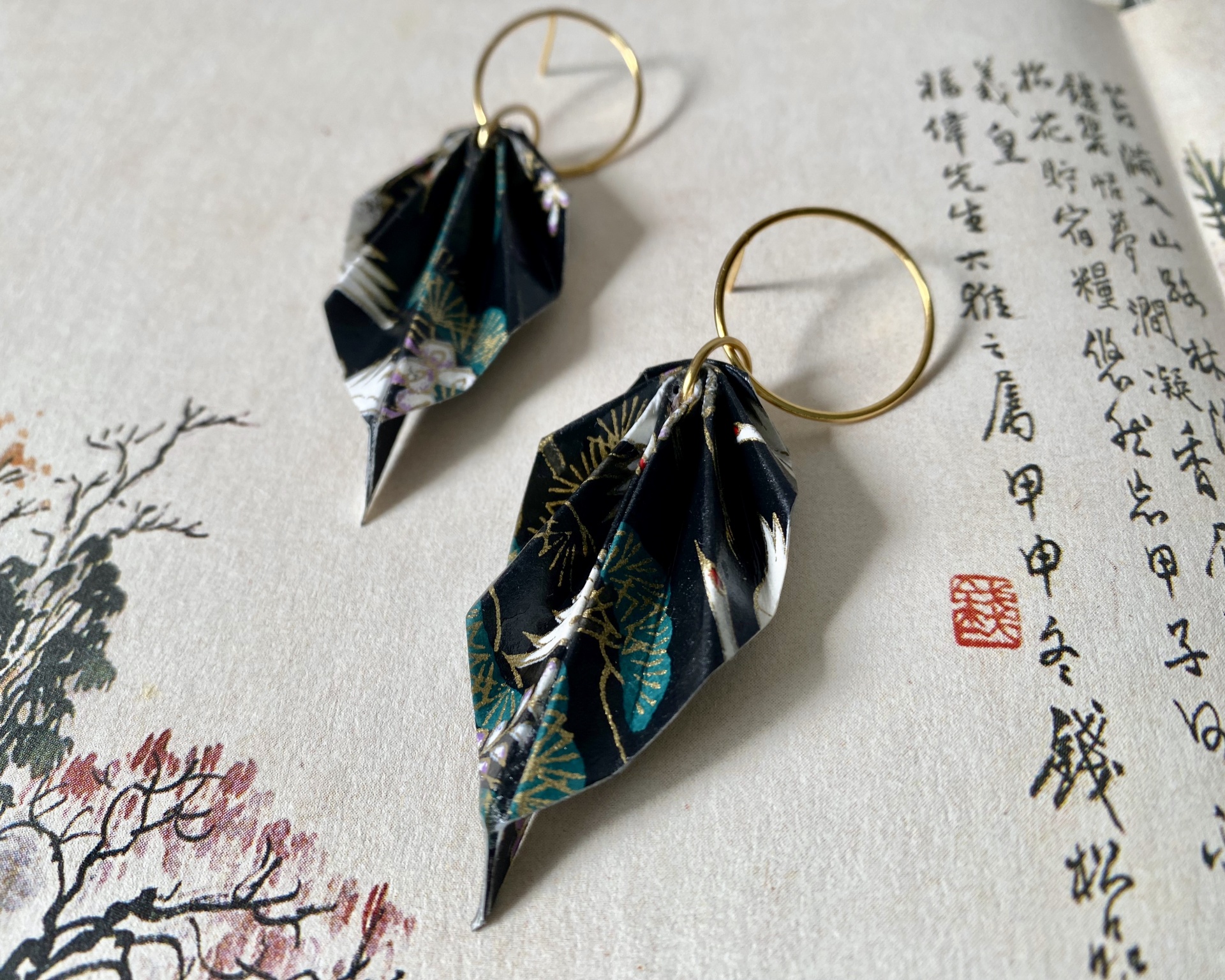 Brincos Folha Brincos pendentes de papel floral preto com argolas douradas sobre papel com caligrafia asiática.