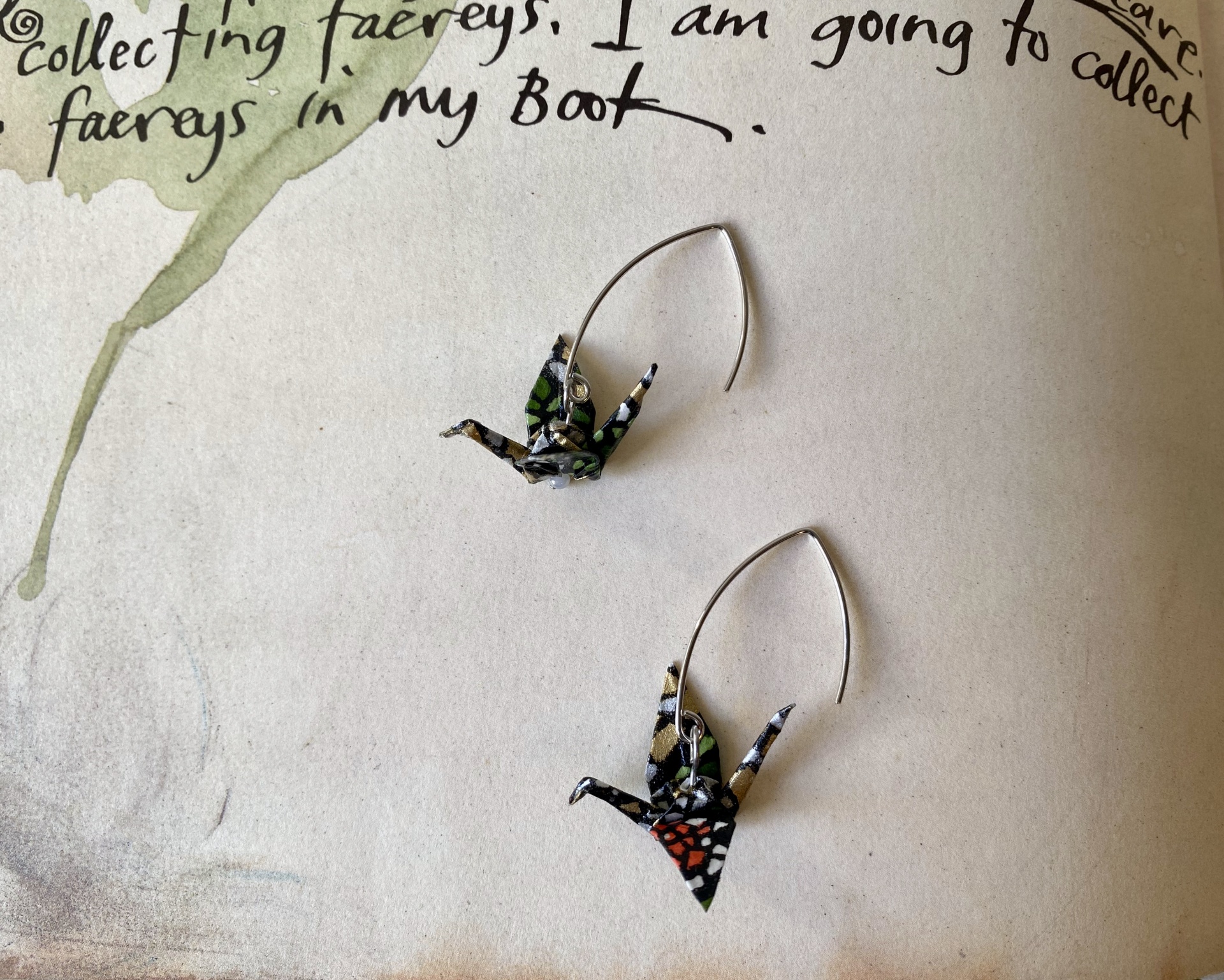 Brincos Tsuru Brincos com origami de pássaros coloridos e base metálica prateada sobre papel texturado com escrita preta.