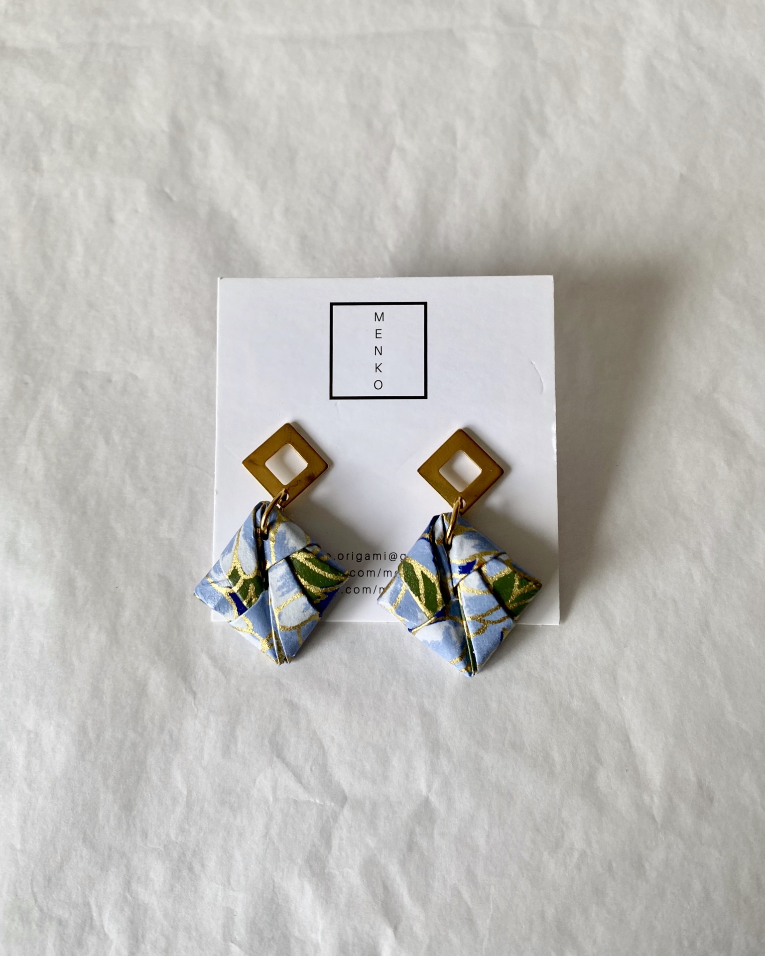 Brincos Sonobe Brincos com base quadrada dourada e peça em origami azul claro com padrões verdes e dourados no cartão MENKO.