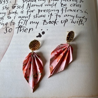 Brincos com base dourada e pendente de papel rosa com padrão floral, sobre página de livro manuscrito.