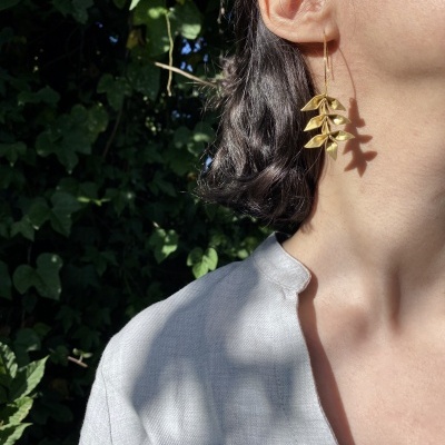 Brincos dourados em formato de folhas usados por pessoa com camisa cinza claro em ambiente com vegetação verde