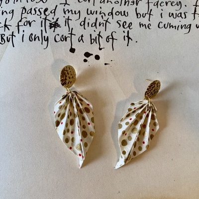 Brincos com tecido branco e bolinhas douradas e vermelhas sobre papel manuscrito