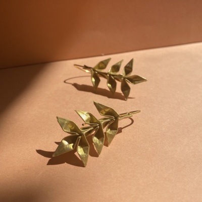 Brincos dourados em forma de raminho com folhas, sobre fundo castanho claro.
