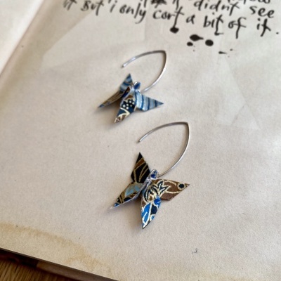 Par de brincos em forma de borboleta com padrões azuis e castanhos sobre uma página de livro