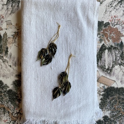 Brincos dourados com folhas negras sobre pano branco com franjas em superfície decorada