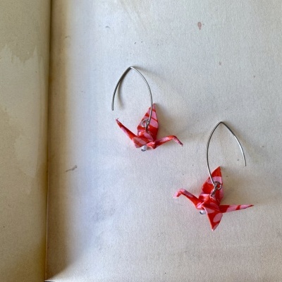 Brincos origami em forma de pássaros vermelhos em fundo branco ao lado de página de livro com escrita manuscrita.