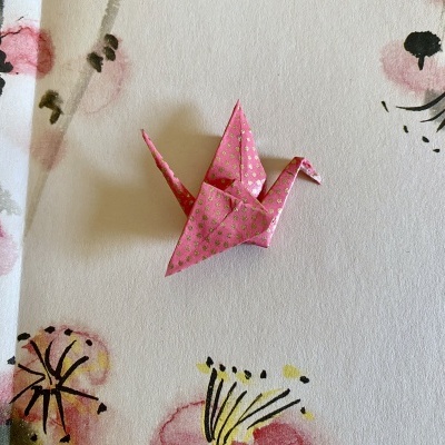 Origami de pássaro rosa com padrão dourado sobre fundo branco com flores pintadas.
