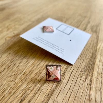 Brinco quadrado em metal com padrão de tecido rosa e flores brancas sobre cartão branco em superfície de madeira clara.