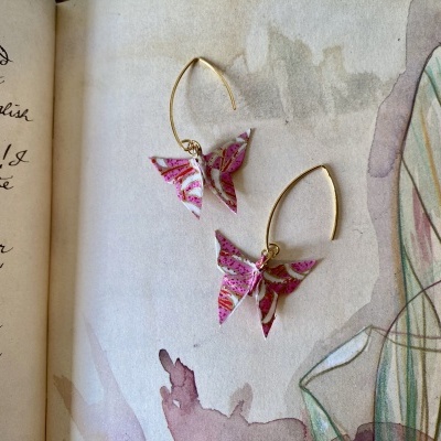 Brincos de borboleta rosa e branco com arame dourado sobre livro aberto.