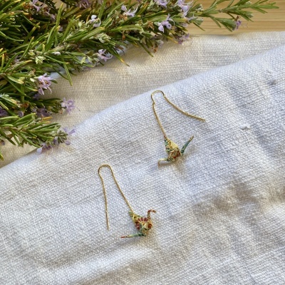 Brincos pendentes com pássaros origami coloridos sobre tecido branco com ramos de flores lilases