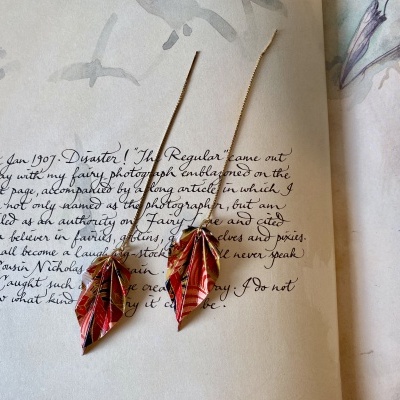 Brincos dourados com folhas vermelhas sobre página manuscrita