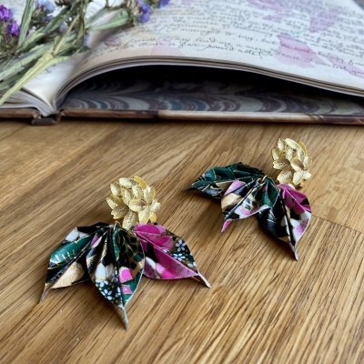 Brincos de origami coloridos com fechos dourados sobre madeira com caderno aberto e flores secas