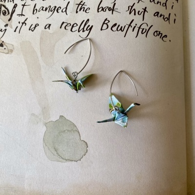 Brincos de origami em forma de crane em papel azul e verde com argolas prateadas sobre papel manuscrito