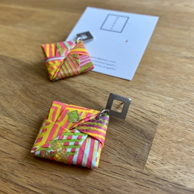 Brincos quadrados coloridos com padrões geométricos e detalhes metálicos sobre mesa de madeira clara