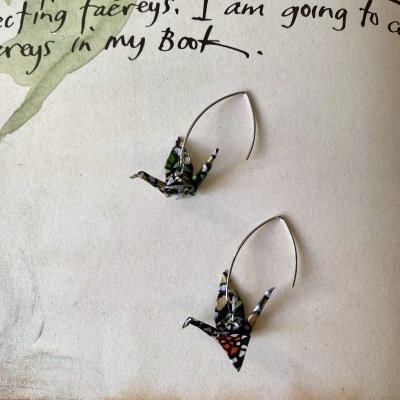 Brincos com origami de pássaros coloridos e base metálica prateada sobre papel texturado com escrita preta.