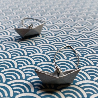 Brincos em forma de barco de papel prateados num fundo com padrão de ondas azuis e brancas
