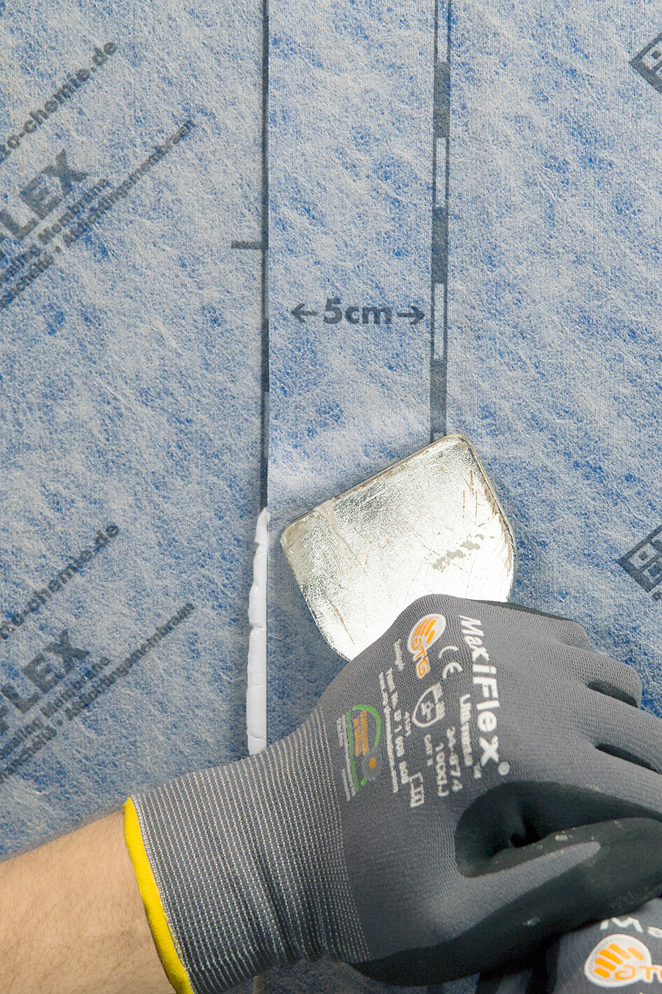 OTTOCOLL® M 595 Mão com luva cinzenta segurando espátula sobre manta azul claro com palavras e marca MacFlex.