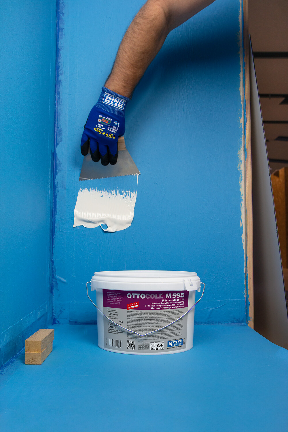 OTTOCOLL® M 595 Balde OTTOCOLL M 595 com massa branca aplicado na parede azul com espátula e mão enluvada