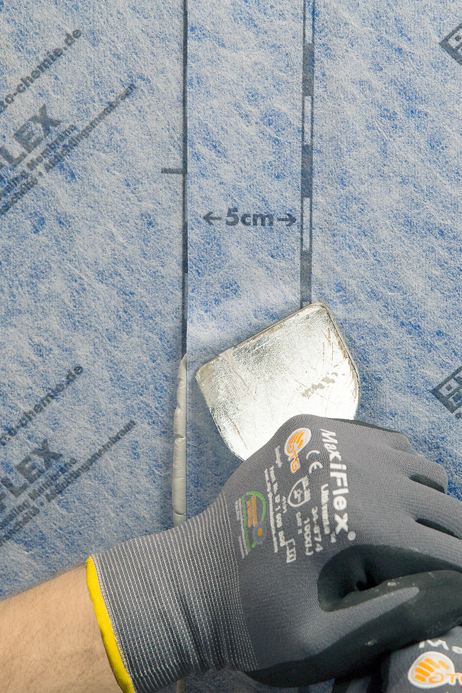 OTTOFLEX® - Tela de Impermeabilização Mão com luva segurando espátula sobre superfície azul com texto Maliflex e marcação 5cm