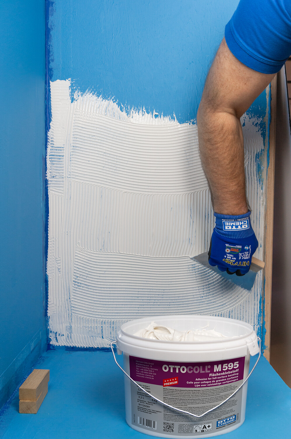 OTTOCOLL® M 595 Balde de adesivo OTTICOLL M 595 no chão azul com pessoa a aplicar adesivo na parede azul
