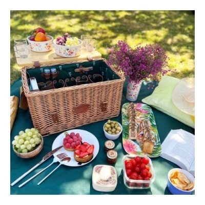 Cesta de piquenique de vime com ferramentas, frutas, flores e mantas num piquenique ao ar livre