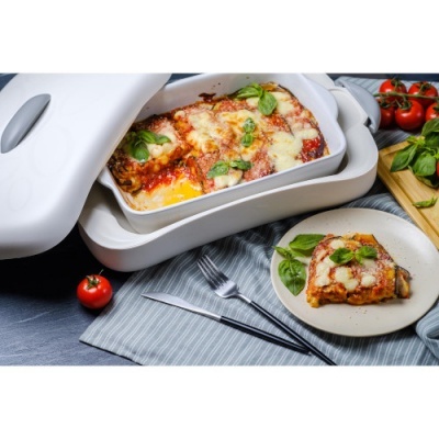 Caçarola branca com lasanha numa base aquecida bege, talheres e tomate cherry ao lado