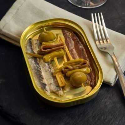 Lata aberta de conservas de peixes com azeitonas e pimentos sobre pano cinza e garfo