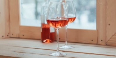 Duas taças com vinho rosé e uma garrafa sobre mesa de madeira junto a janela