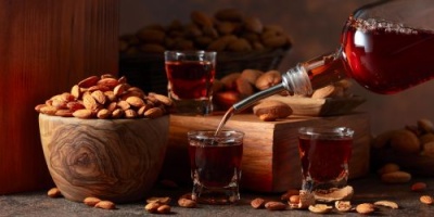 Copos com licor vermelho a ser servido, amêndoas em taça de madeira e cesta ao fundo