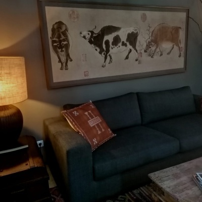 Sofá cinza escuro com almofadas terracota, mesa de madeira, luminária com abajur bege e quadro com vacas em parede abobadada