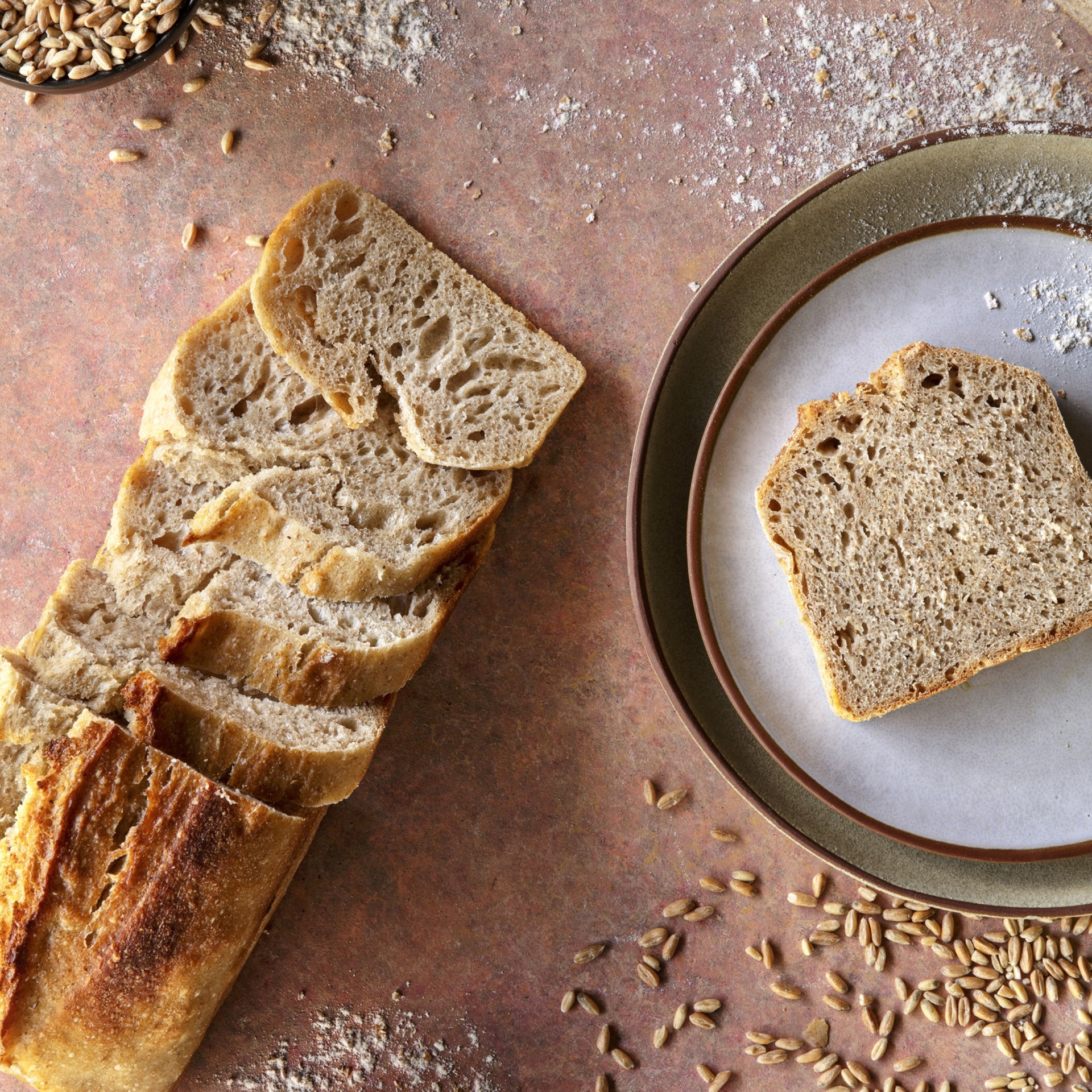 Pão de Espelta Integral BIO 450g - Pachamama Fatias de pão integral e uma fatia no prato sobre superfície com farinha e grãos