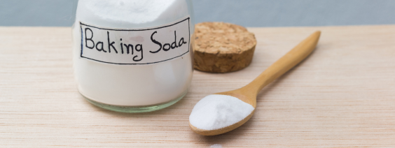 As Utilidades do Bicarbonato de Sódio Frasco de vidro com pó branco e etiqueta Baking Soda ao lado de colher de madeira com pó branco.