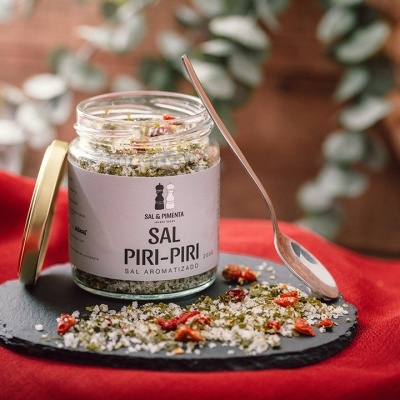Frasco de sal aromatizado Sal Piri-Piri com tampa dourada e sal temperado espalhado em prato de ardósia